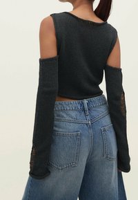Woman wearing a dark gray sleeveless knit top with detached long sleeves and high-waisted blue denim jeans, shown from the back.
