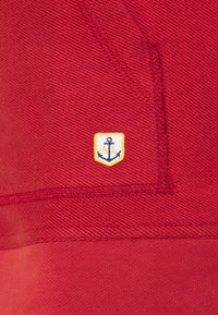 Sudadera roja hecha de tejido texturizado que presenta un pequeño parche bordado en forma de escudo con un ancla y "AL" en azul y amarillo.
