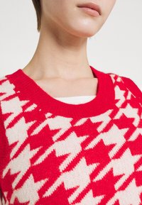 Czerwono-biały sweter w kratę pepitkę z szerokim dekoltem i prążkowanym kołnierzem. Materiał wydaje się miękki i dziany, ukazując wyraźną fakturę.