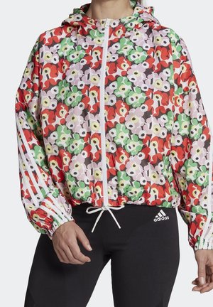 Kurtka z kapturem na zamek błyskawiczny w kwiatowy wzór z czerwonymi, zielonymi i białymi kwiatami, białe paski na rękawach, noszona z czarnymi legginsami Adidas.