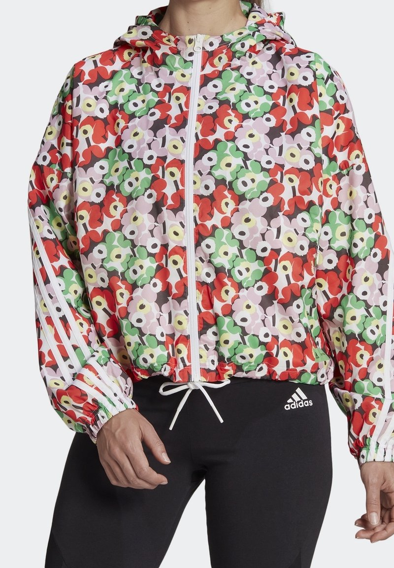 Veste à capuche zippée à motif floral avec des fleurs rouges, vertes et blanches, bandes blanches sur les manches, portée avec des leggings noirs Adidas.