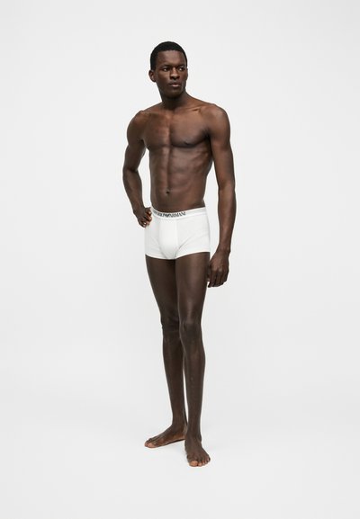 Γυμνασμένος άνδρας με σκούρο δέρμα, όρθιος ας ξυπόλητος, φοράει λευκά μποξεράκια Armani, με απλό ανοιχτό γκρι φόντο, κοιτάζει στο πλάι.
