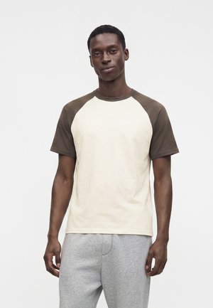 Homme portant une chemise beige à manches courtes avec des manches marron foncé et un pantalon de survêtement gris, debout devant un fond blanc uni.