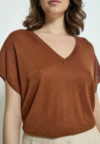 Top de punto con escote en V en color óxido con brillos metálicos, manga corta y un corte relajado, combinado con pantalones de color claro.