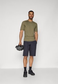 Chemise olive à manches courtes, short noir, chaussures noires, tenant un casque de vélo noir. Design simple, texture lisse, style fonctionnel.