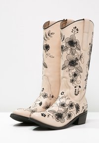 Stivali da cowboy in pelle beige con elaborate ricamate floreali neri. Punta quadrata, tacco basso e gambale appuntito con cuciture decorative.