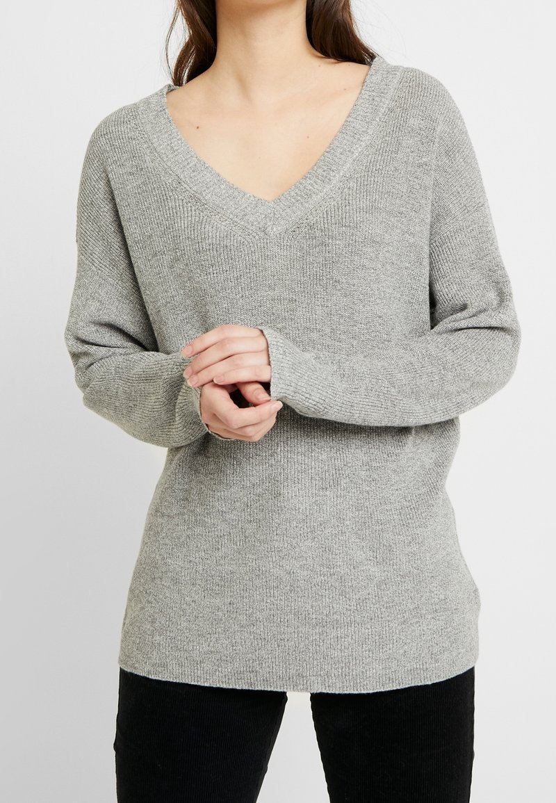 Femme portant un pull en maille gris clair à col en V avec des manches longues et un pantalon noir, debout devant un fond blanc uni.