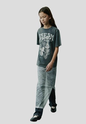 Grå grafisk t-shirt med vit "FRESH" text; lager med en genomskinlig spetskjol; mörka byxor och svarta skor med en glänsande finish.