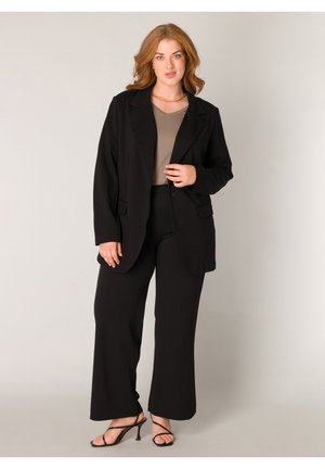 Frau mit hellbraunen Haaren, die einen schwarzen Blazer, schwarze weite Hosen, ein taupefarbenes Oberteil und schwarze Riemchenabsatzsandalen trägt, steht vor einem hellen Hintergrund.