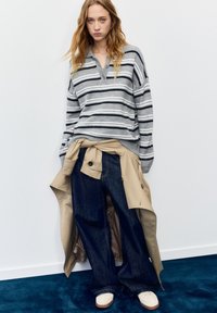 Grijs-wit gestreepte gebreide trui met een kraag, gecombineerd met wijde donkerblauwe spijkerbroeken. Een beige trenchcoat gestrikt in de taille. Witte schoenen.