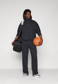 Sudadera negra con bolsillo frontal y logo, combinada con pantalones a juego. Sosteniendo un balón de baloncesto naranja y una bolsa de gimnasio negra con una correa de logo.