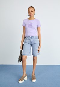 Light purple short-sleeve t-shirt with logo, blue denim shorts, and beige platform sandals with a white sole, holding a brown handbag.