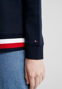 Felpa blu navy con polsini a costine e un orlo colorato in bianco e rosso. Presenta un piccolo logo vicino al polsino.