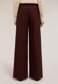 Osoba v tmavě hnědých kalhotách s vysokým pasem a širokými nohavicemi a béžovém pleteném svetru, stojící proti jednolité světlé pozadí.