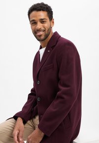 Bordeauxkleurige wolmixjas met ingesneden revers, drie knopen, zijzakken en een logo detail op de borstzak. Model draagt een licht shirt en een broek.