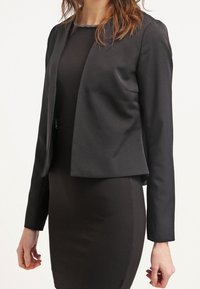 Svart kort blazer i slätt tyg, med rund halsringning och långa ärmar, kombinerad med en åtsittande svart klänning.