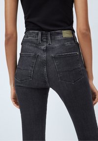 Kvinna som bär högmidjade, mörkgrå denimjeans och en svart topp, sedd från ryggen i midjehöjd mot en enfärgad bakgrund.