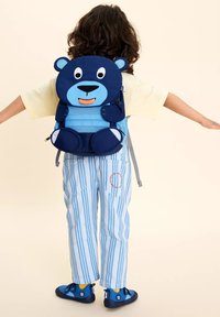 Blauer Bären-Rucksack mit runden Ohren und Gesicht, das ein Lächeln zeigt. Gestreifte blaue Hose mit roter Stickerei. Rückansicht eines Kindes.