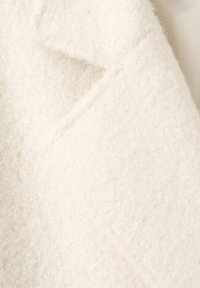 Abrigo blanco de mezcla de lana con un acabado suave y texturizado. Presenta un cuello con muescas y detalles de costura visibles. La vista en primer plano muestra su superficie difusa.