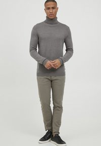 Grau gestrickter Rollkragenpullover aus Strickstoff, körperbetont. Kombiniert mit hellgrauen Hosen und schwarzen Sneakers, vor einem schlichten Hintergrund stehend.