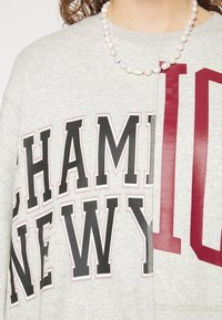Felpa grigia con grande scritta nera e bordeaux che recita "CHAMPION NEW YORK." Presenta un design a spacco e un tessuto leggero.