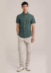 Camisa de botón de manga corta verde con una textura sutil, combinada con pantalones beige claros y zapatillas blancas con suelas de goma.