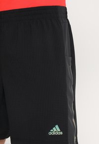 Pantalones cortos deportivos negros con textura acanalada, con una cintura elástica, bolsillos laterales y un logo verde de Adidas.
