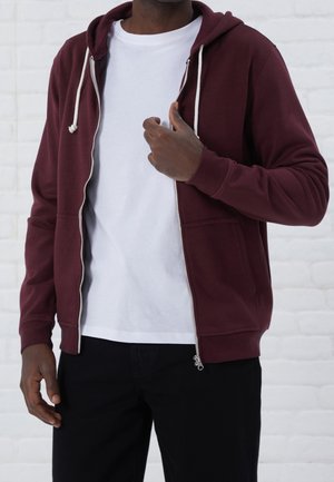 Uomo che indossa una felpa con cappuccio bordeaux con zip sopra una t-shirt bianca e pantaloni neri, aggiustando la cerniera con una mano.