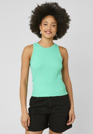 Femme aux cheveux bouclés portant un haut sans manches vert menthe et un short noir, souriante sur un fond uni gris clair.