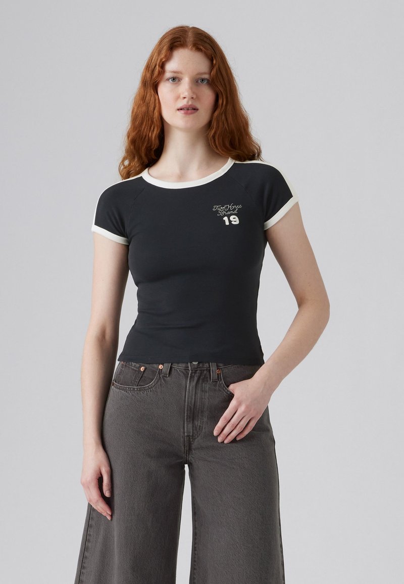 T-shirt noir ajusté avec des manches raglan courtes, bord blanc, et un texte graphique à l'avant indiquant "True Horse Brand 19." Associé à un jean gris à jambes larges.
