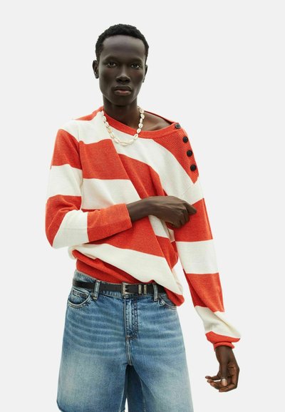 Gestreifter Pullover in rot und weiß, aus strukturiertem Strickstoff, mit schulterfreiem Design und schwarzen Knöpfen an einer Schulter; kombiniert mit Jeansshorts.