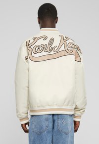 Cremfarbener Bomberjacke mit großen beigen "Karl"-Buchstaben auf dem Rücken, gerippten Bündchen und Saum sowie glatter satinähnlicher Textur. Das Model trägt hellblaue Jeans.