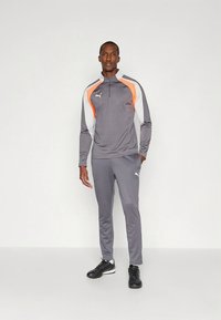 Szürke sportos melegítő narancssárga és fehér részletekkel, cipzáras nyakkal, hosszú ujjakkal és testhez simuló nadrággal. Fekete sportcipő teszi teljessé a megjelenést.