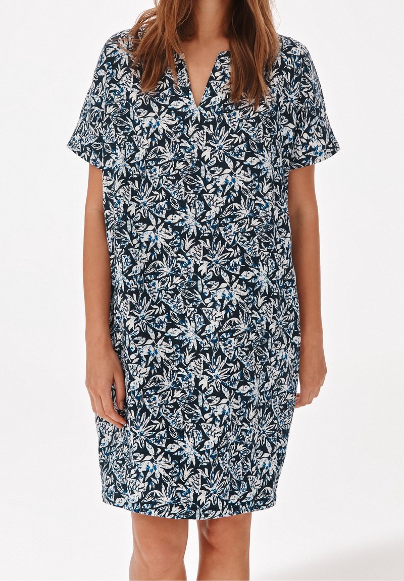 Femme portant une robe à manches courtes, longueur au genou, avec un motif floral bleu marine, blanc et bleu, et un col en V.