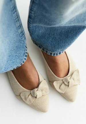 Pies calzando bailarinas puntiagudas texturizadas en beige con lazos de tela, combinadas con jeans de mezclilla azul claro sobre un fondo blanco.