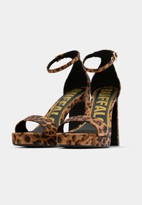 Sandalias de tacón alto con plataforma y estampado de leopardo, con tira en el tobillo y suela exterior negra. Logo amarillo visible en la plantilla. Textura de tela suave.