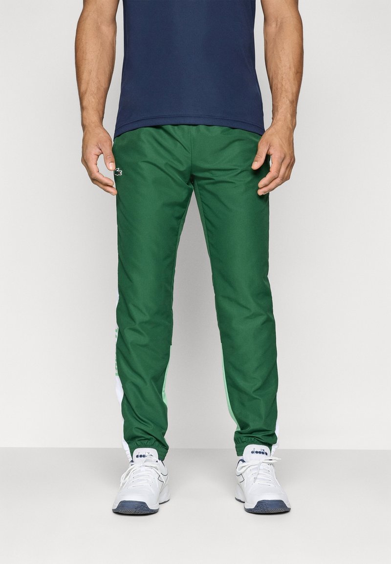 Pantalones deportivos verdes con cinturilla elástica, franjas blancas y verdes a los lados, y logotipo bordado. Combinados con zapatillas blancas.