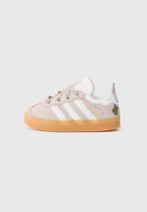 Light gray suede sneaker with white stripes, rubber gum sole, padded collar, and a decorative patch on the heel featuring a cartoon design.