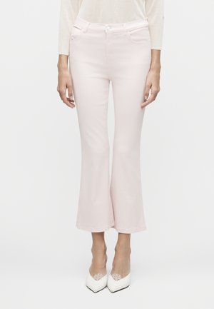 Γυναίκα που φοράει ανοιχτό ροζ ψηλόμεσα παντελόνια καμπάνα με κοντό μήκος, συνδυασμένα με λευκές μυτερές γόβες με δαντελένιες λεπτομέρειες.