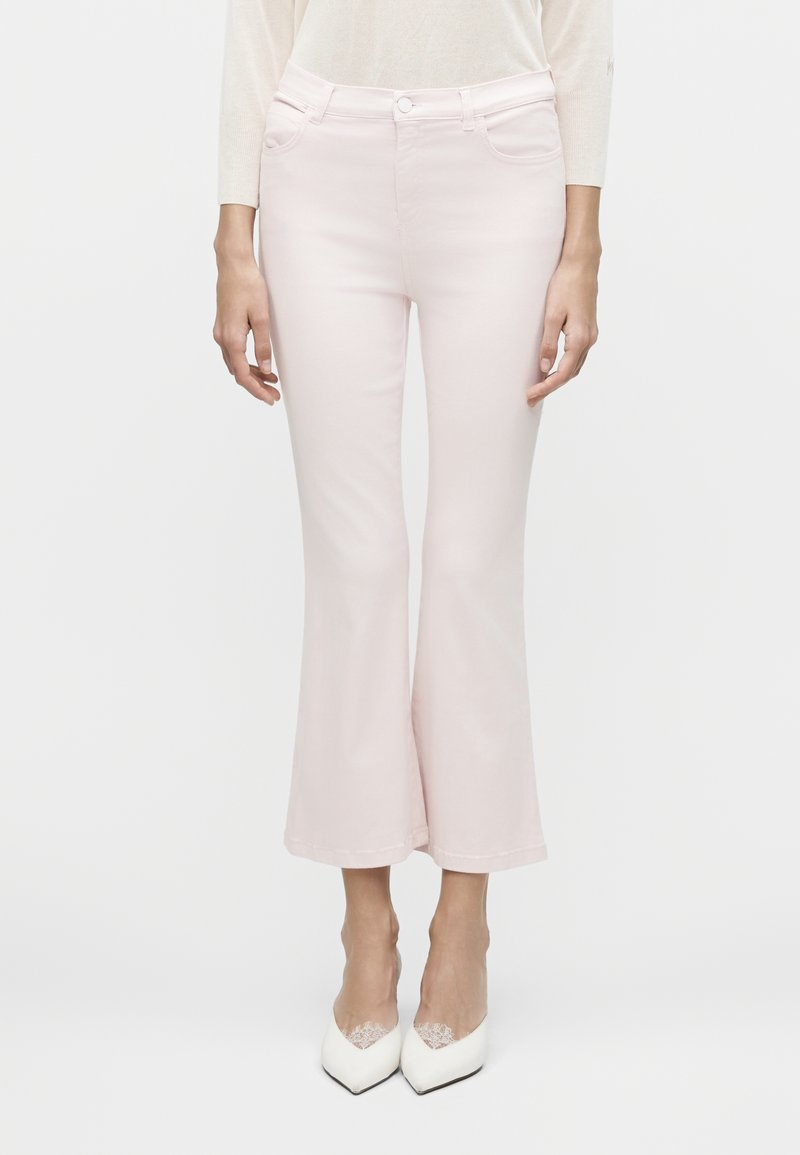 Γυναίκα που φοράει ανοιχτό ροζ ψηλόμεσα παντελόνια καμπάνα με κοντό μήκος, συνδυασμένα με λευκές μυτερές γόβες με δαντελένιες λεπτομέρειες.