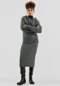 Woman with curly hair wears grey knit hoodie and matching skirt with black turtleneck and knee-high black boots, standing with arms crossed.