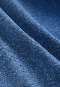 Blauer Denimstoff mit diagonalen Falten, der eine detaillierte gewebte Textur und leichte Farbvariationen zeigt, die für Jeansmaterial typisch sind.