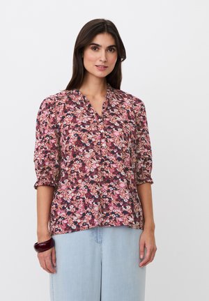 Mujer con una blusa rosa con flores y mangas tres cuartos y pantalones de talle alto azul claro, de pie contra un fondo blanco liso.