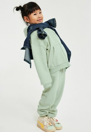 Enfant avec deux petits chignons portant un survêtement vert clair, des baskets beiges et une veste en jean posée sur les épaules, se penchant légèrement en arrière.