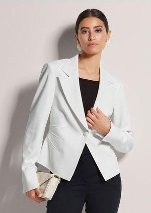 Frau mit dunklen Haaren, die einen weißen Blazer über einem schwarzen Top und Hosen trägt, hält eine beige Clutch und steht vor hellem Hintergrund.
