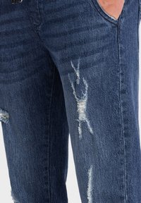 Jeans de mezclilla azul oscuro con un diseño desgastado que presenta parches deshilachados y decoloración. Corte clásico con bolsillos visibles a los lados.