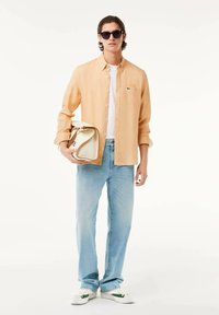 Camicia button-up arancione chiaro sopra una t-shirt bianca, abbinata a jeans blu sbiaditi e sneakers bianche con dettagli verdi. Porta una borsa color crema.