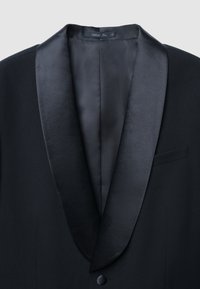 Giacca da smoking nera con rever a scialle in raso. Presenta un solo bottone e una tasca, mostrando un interno in tessuto liscio e strutturato.