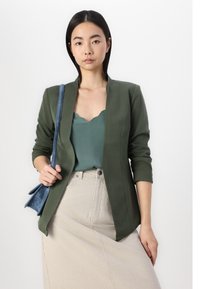 Groene getailleerde blazer met een geschulpt kraag, gecombineerd met een teal top met een scalloped halslijn en een beige geribbelde rok. Model houdt een kleine blauwe tas vast.