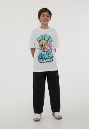 T-shirt bianca con un design grafico colorato che presenta elementi da fumetto e testo sul retro, abbinata a pantaloni neri e sneakers beige.